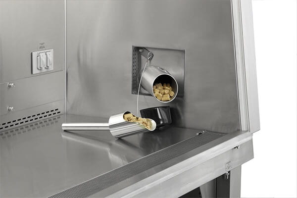 stainless steel biosafety cabinet interior with built-in feed hopper dispensing animal chow onto scoop, designed for efficient vivarium cage changing workflow
